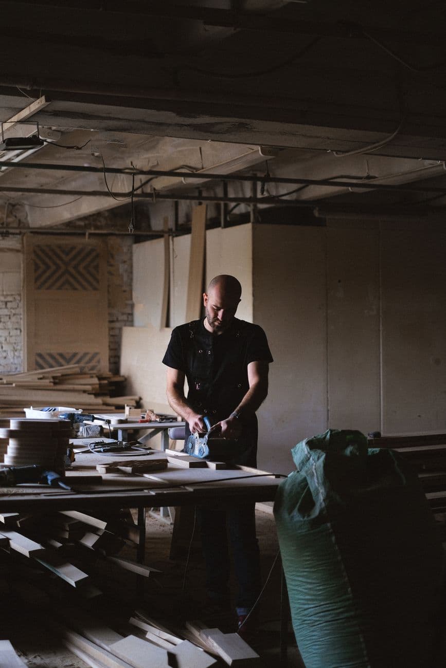 Concentrated male carpenter using sander to polish wooden plank while working at table in professional joinery with stack of planks